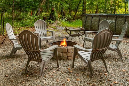 Fire pit surrounded by six adirondack chairs with flames burning at dusk in a wooded clearing.