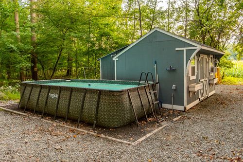 Rectangular above-ground pool with a ladder beside a blue storage shed in a wooded yard.