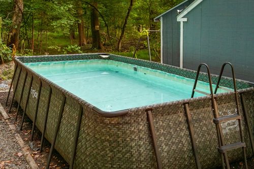 Above-ground woven-panel pool with a ladder, set beside a wooded backyard and storage shed.