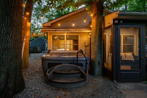 Hot tub with pull-out steps set on a gravel patio beside the cabin, lit by string lights at dusk.