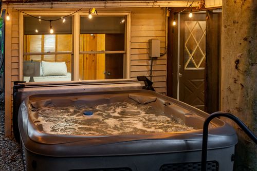Bubbling hot tub on a gravel patio at night, with a lit bedroom visible through the window behind it.