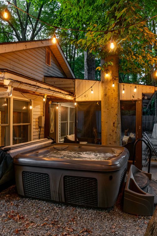 Hot tub on a stone patio beneath string lights, with a screened porch glowing behind it at dusk.