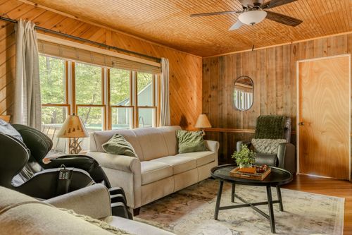 Living room with knotty pine walls, a round mirror and a large picture window framing the woods.