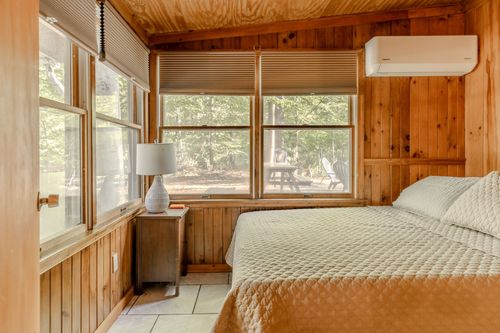 Wood-paneled bedroom with a wall-mounted AC unit and windows overlooking the woods.