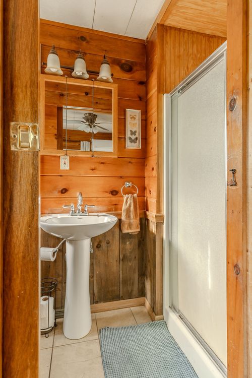 Bathroom with a white pedestal sink, glass shower stall and knotty pine walls.