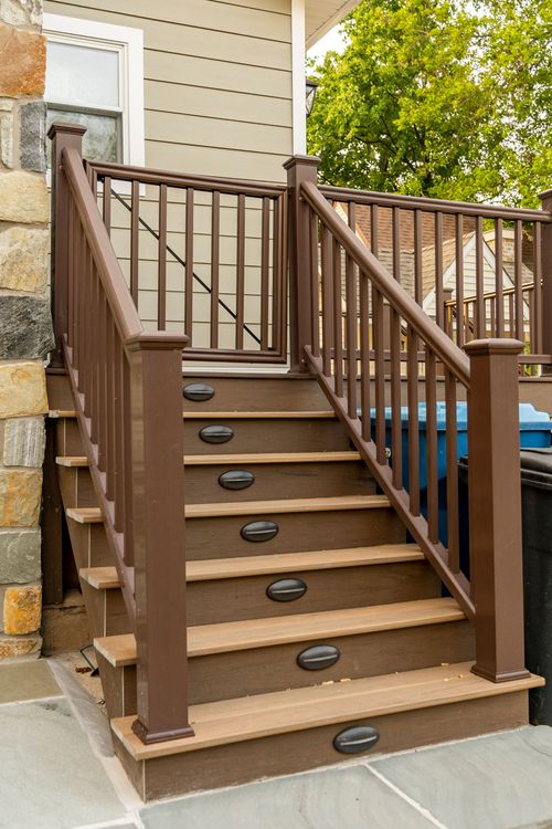 Deck staircase with dark wood railings and round tread lights beside a stone column