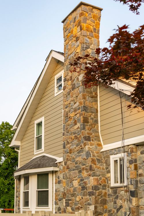 Close view of a tall stone chimney rising against a tan siding gable and red maple tree
