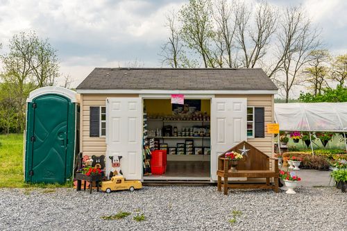 Garden shed with open double doors displaying jars and toys beside a green portable restroom