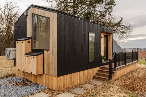Black-and-cedar tiny home exterior with covered deck and staircase to a gravel path