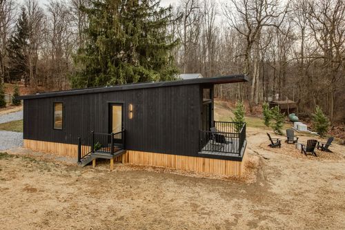 Black tiny cabin with covered deck and Adirondack chair seating around a fire pit clearing
