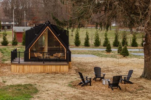 Black A-frame cabin lit with string lights beside deck seating and a nearby fire pit at dusk