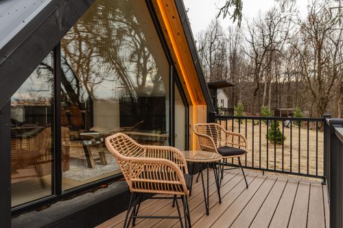 Deck corner with wicker chairs beside an A-frame cabin's glass wall reflecting bare trees