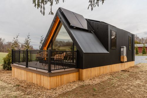 Black A-frame cabin with solar-panel roof and an extended rectangular wing over cedar skirting