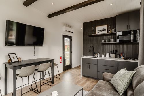 Open living space with beam ceiling, mounted TV, dining table and a dark galley kitchenette