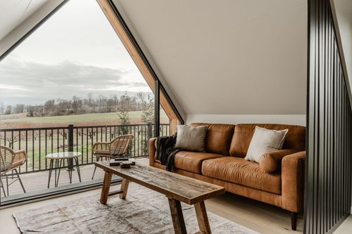 Tan leather sofa and reclaimed wood bench beneath an A-frame window overlooking farmland