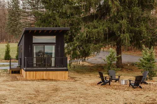Black tiny cabin nestled among evergreens with a covered deck and angular roofline