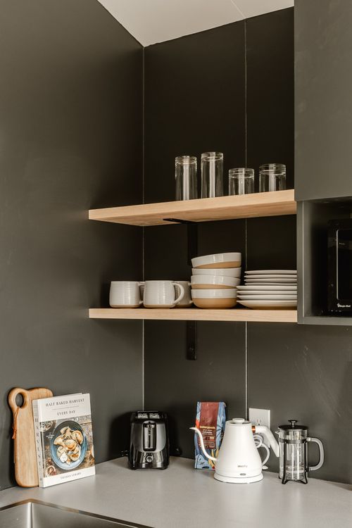 Dark kitchenette open shelving with glassware, stacked bowls, a cookbook and kettle