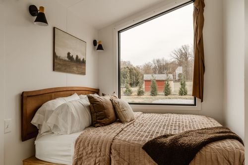 Bedroom with a cane headboard, layered brown pillows and a window overlooking a red barn
