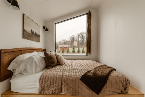 Bedroom with cane headboard, layered pillows and a tan bedspread beside a farmland window