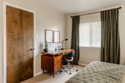 Bedroom corner with a wood desk, task lamp and olive curtains beside a wood door