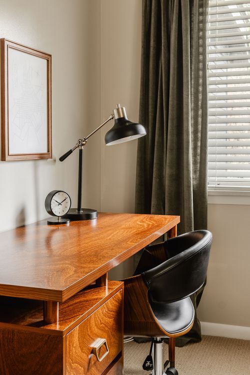 Wood desk with task lamp and clock beside olive curtains and a window