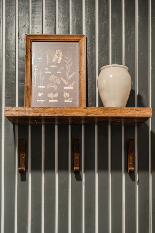 Framed botanical print and ceramic vase on a wood shelf against a dark slat wall