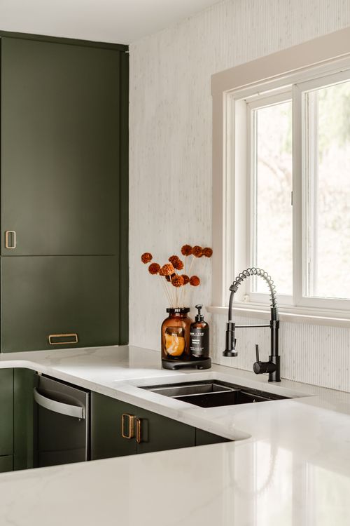 Kitchen corner with green cabinets, brass fixtures and a vase of dried flowers by a window