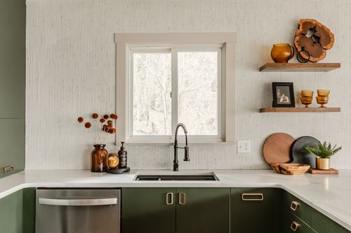 Kitchen sink under a window with green cabinetry and open shelves of amber glassware