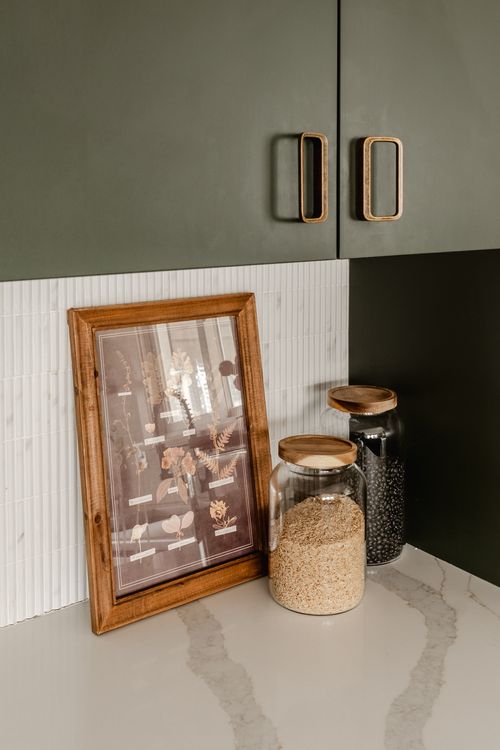 Glass storage jars and a framed botanical print on a marble counter beside green cabinets