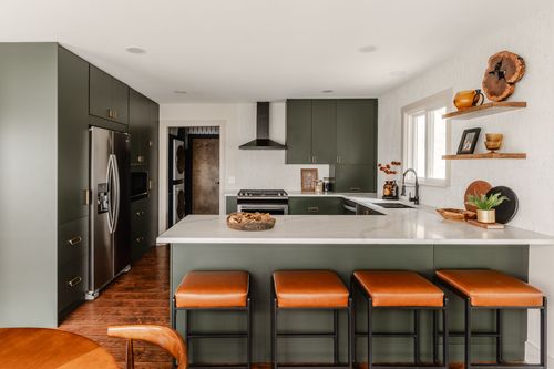 Kitchen with dark green cabinetry, stainless fridge, black hood vent and tan leather counter stools