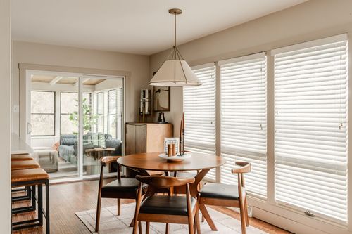 Round wood dining table with black leather chairs beneath tall windows with white blinds