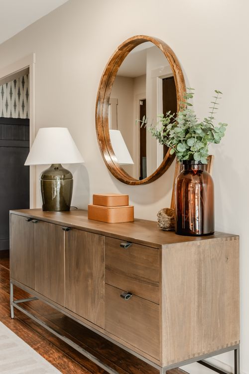 Round wood mirror above a wood console table styled with a lamp and leather boxes