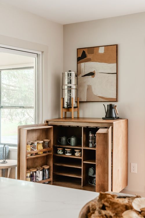 Open wood coffee cabinet with mugs, jars and a French press beneath abstract wall art