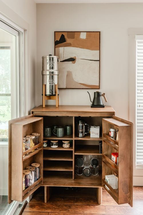 Open wood cabinet styled as a coffee bar with mugs, French press and storage jars