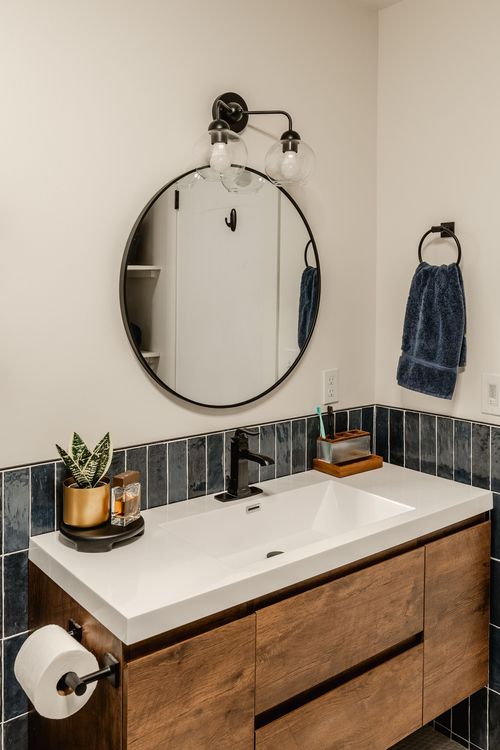 Round mirror and glass sconce over a navy tile bathroom with a wood vanity and towel ring