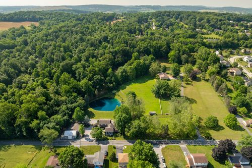 Aerial view of a wooded property with a round pond and open lawn beside a country road