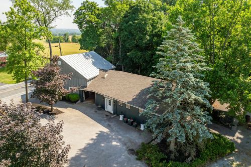 Aerial view of a metal-roofed outbuilding beside a gravel driveway and blue spruce tree
