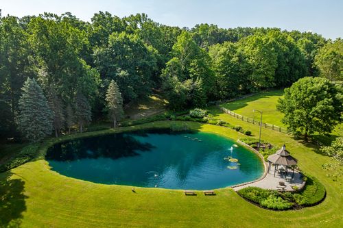 Aerial view of a curved pond bordered by manicured lawn, a gazebo and mature trees