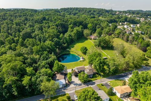 Aerial view of a farmhouse-style property with a pond, set among mature trees near a residential road