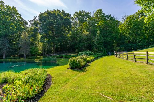 Green pond edged with split-rail fencing, ornamental grasses and a footbridge in the distance