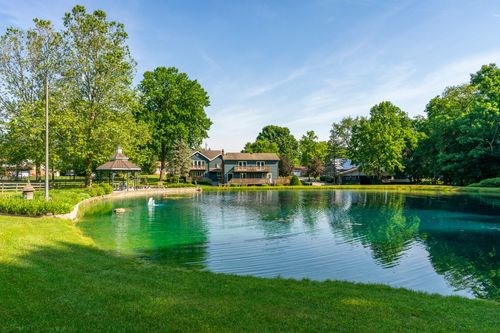 Large home reflected in a green pond with a gazebo and mature trees along the shore