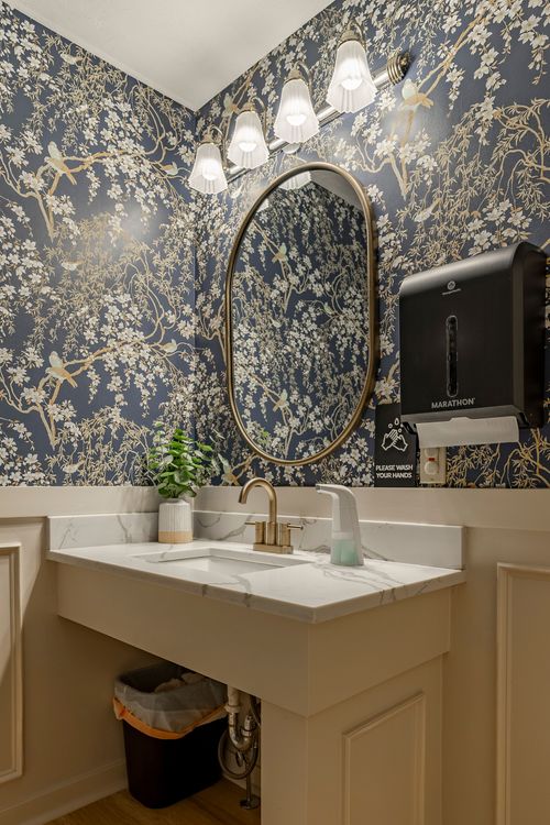 Close view of a marble vanity beneath an oval brass mirror and navy bird-print wallpaper
