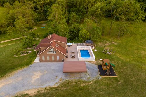 Aerial view of a farmhouse with a pool and fire pit patio bordered by woodland