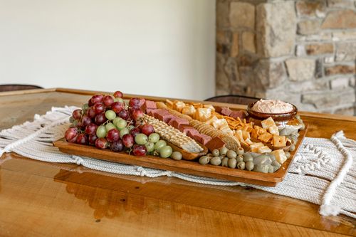 Wood charcuterie tray with grapes, corn and cured meats beside a stone fireplace