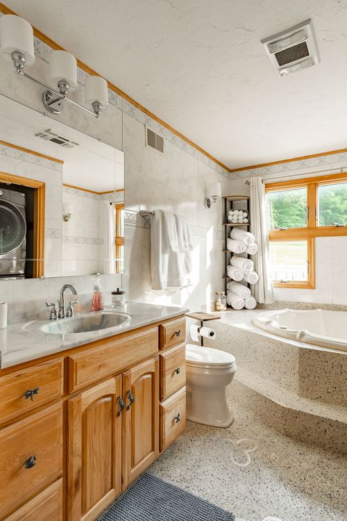 Bathroom with an oak vanity, terrazzo flooring, tiled walls and a large jetted tub by a window