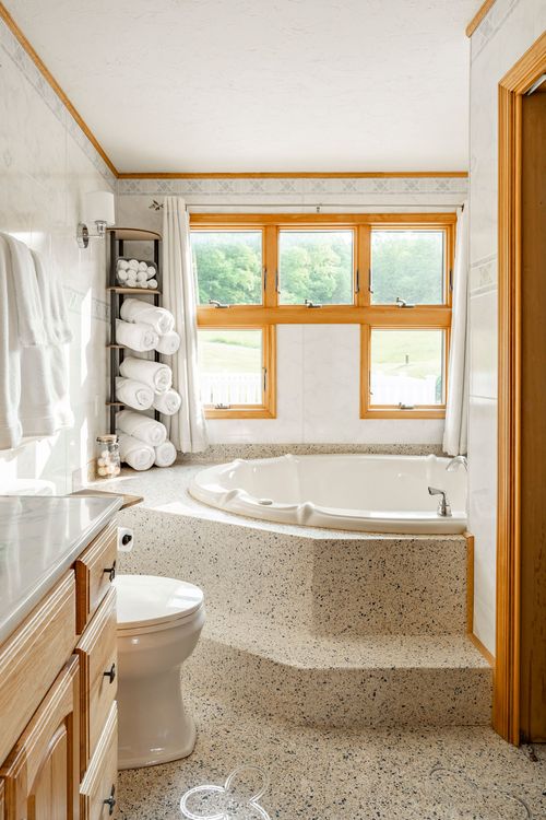 Corner jetted tub beneath a triple window with a terrazzo tile deck and warm wood trim