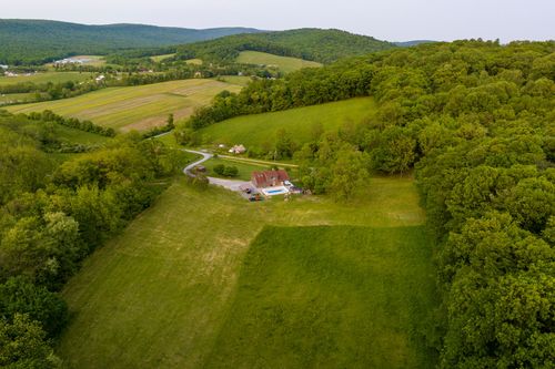 Aerial view of a farmhouse with a pool along a curving driveway framed by wooded hills