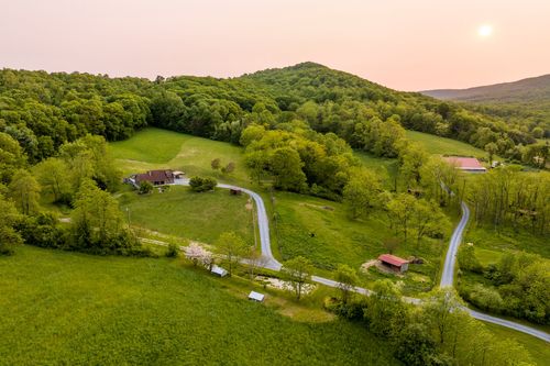 Aerial sunset view of a farmhouse nestled in green pasture beneath a wooded ridge