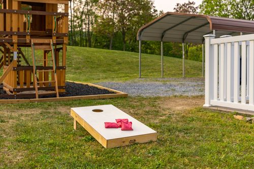 Cornhole board set on grass near a play structure and carport