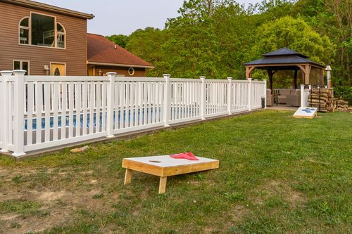 Cornhole board on the lawn beside a white picket pool fence and gazebo